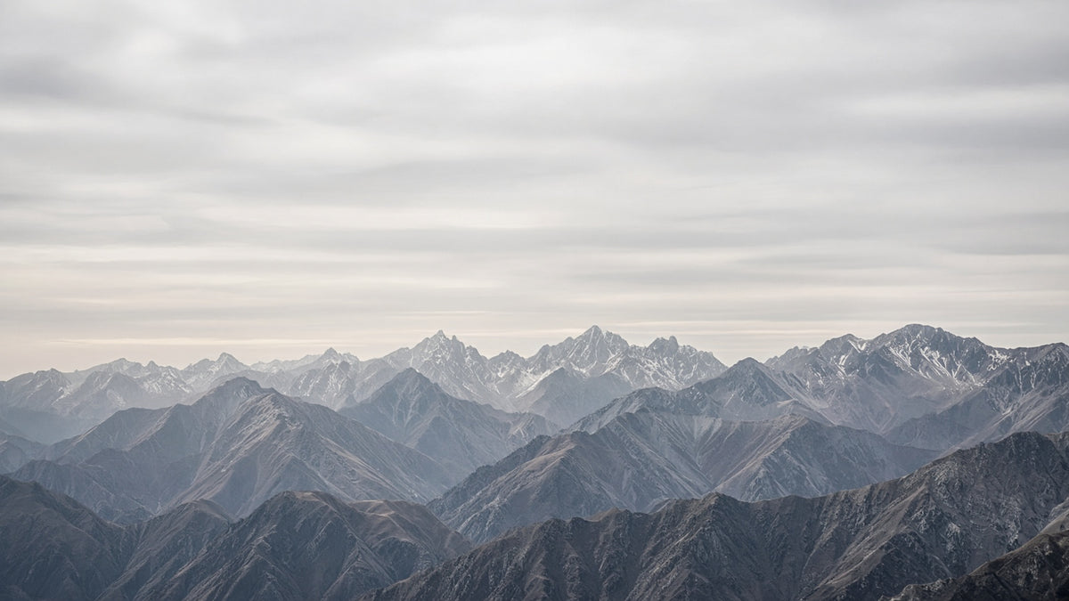 A scenic landscape photograph of layered mountain ranges under a cloudy, overcast sky. The mountains show shades of blue, gray, and brown, with some snow-capped peaks visible in the distance.